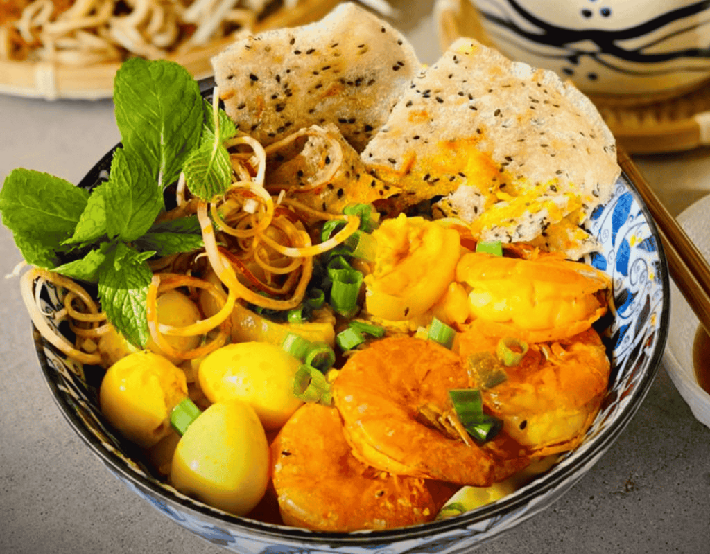 Mi Quang combines turmeric-colored noodles, fresh herbs, savory toppings, peanuts, and crispy rice crackers.